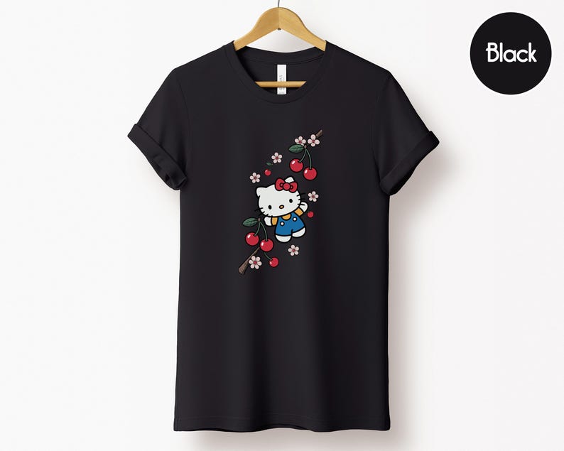 Op de afbeelding: Zwart T-shirt met een cartoonfiguur met een rode strik, blauwe overall en een kersentakontwerp. De kersen zijn rood en de bloemen roze en wit. Het shirt hangt aan een houten hanger.