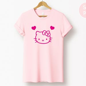 以下が含まれることがあります： 薄いピンク色のTシャツ。マゼンタ色のハローキティのデザインと2つのハートが描かれています。デザインには、ハローキティの顔、リボン、ひげ、目があります。木製のハンガーにTシャツが掛けられています。