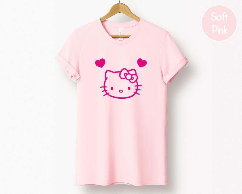 以下が含まれることがあります： 薄いピンク色のTシャツ。マゼンタ色のハローキティのデザインと2つのハートが描かれています。デザインには、ハローキティの顔、リボン、ひげ、目があります。木製のハンガーにTシャツが掛けられています。