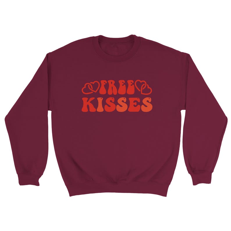 May include: A maroon sweatshirt with long sleeves. The front displays the words "FREE KISSES" in orange and red retro lettering, with heart shapes integrated into the design. This is a casual item of clothing.