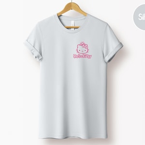 May include: A light gray t-shirt with rolled-up sleeves, hanging on a wooden hanger. The shirt features a pink Hello Kitty graphic on the front. The text "HelloKitty" is below the graphic.