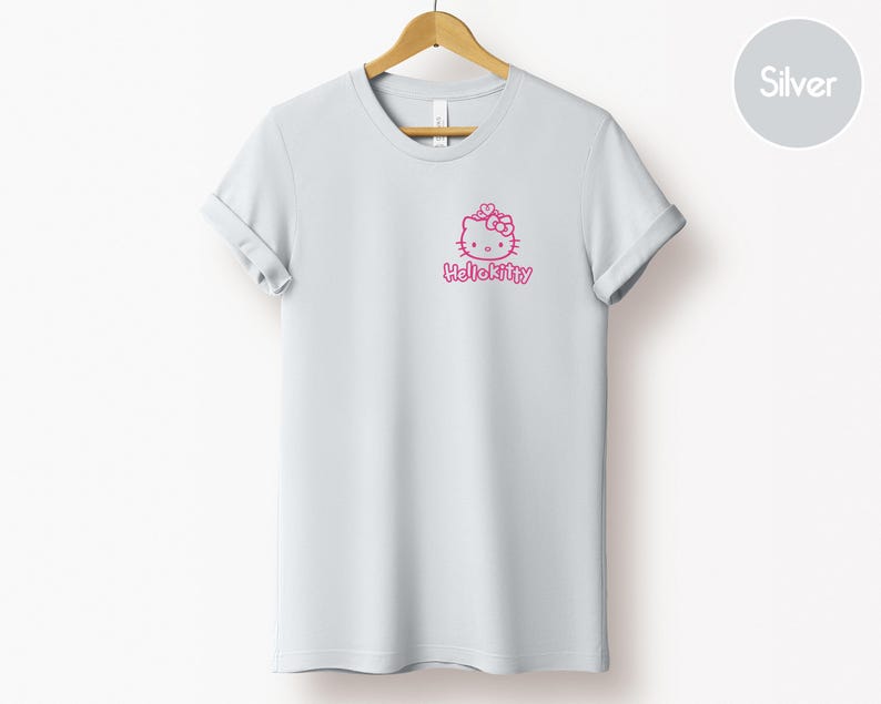 May include: A light gray t-shirt with rolled-up sleeves, hanging on a wooden hanger. The shirt features a pink Hello Kitty graphic on the front. The text "HelloKitty" is below the graphic.
