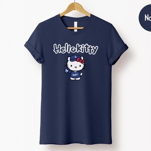 May include: Navy blue t-shirt with a Hello Kitty graphic in white script above a cartoon image of Hello Kitty in a baseball cap and uniform. The shirt is hanging on a wooden hanger against a white background.