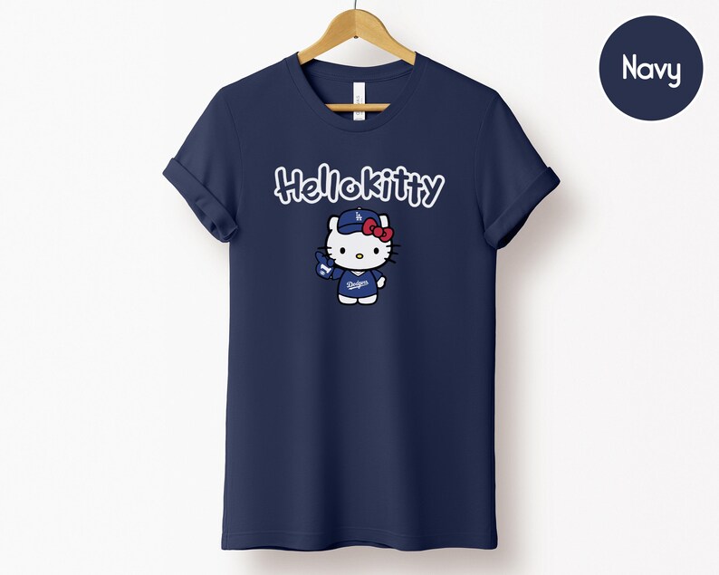 May include: Navy blue t-shirt with a Hello Kitty graphic in white script above a cartoon image of Hello Kitty in a baseball cap and uniform. The shirt is hanging on a wooden hanger against a white background.