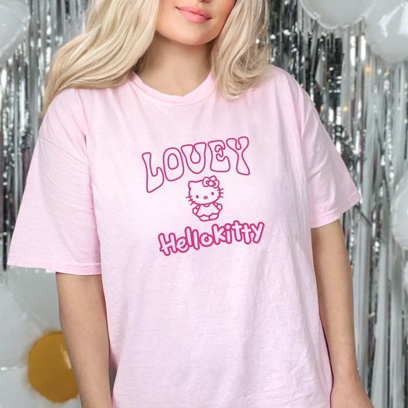 Op de afbeelding: Een lichtroze t-shirt met de woorden "LOVEY" en "HelloKitty" in magenta. Het shirt heeft een Hello Kitty-afbeelding. Het t-shirt is een casual, oversized stijl.