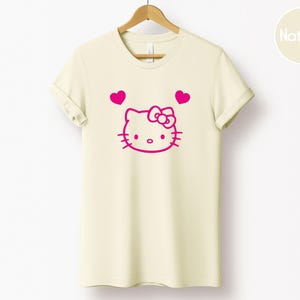以下が含まれることがあります： ピンク色のリボンと2つのピンク色のハートが付いた漫画の猫のピンク色のグラフィックが特徴のクリーム色のTシャツ。 Tシャツは木製のハンガーに吊るされています。