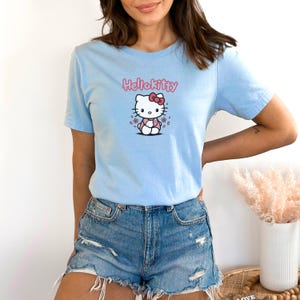 May include: Light blue t-shirt featuring a Hello Kitty graphic. The graphic includes the text "Hello Kitty" in pink above an illustration of the character with a pink bow and flowers. The shirt is short-sleeved.