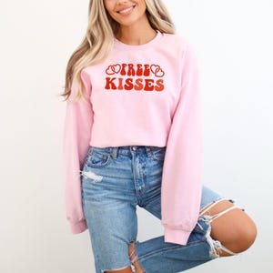May include: A pale pink sweatshirt with long sleeves. The front displays the words "FREE KISSES" in red, with two heart shapes above the word "KISSES". The sweatshirt is worn with blue jeans.