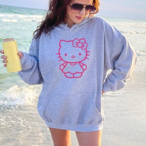 May include: A light gray hoodie featuring a pink outline of Hello Kitty. The hoodie has a front pocket and a drawstring hood. A yellow can cooler is held in the hand. The background is a beach with ocean waves.