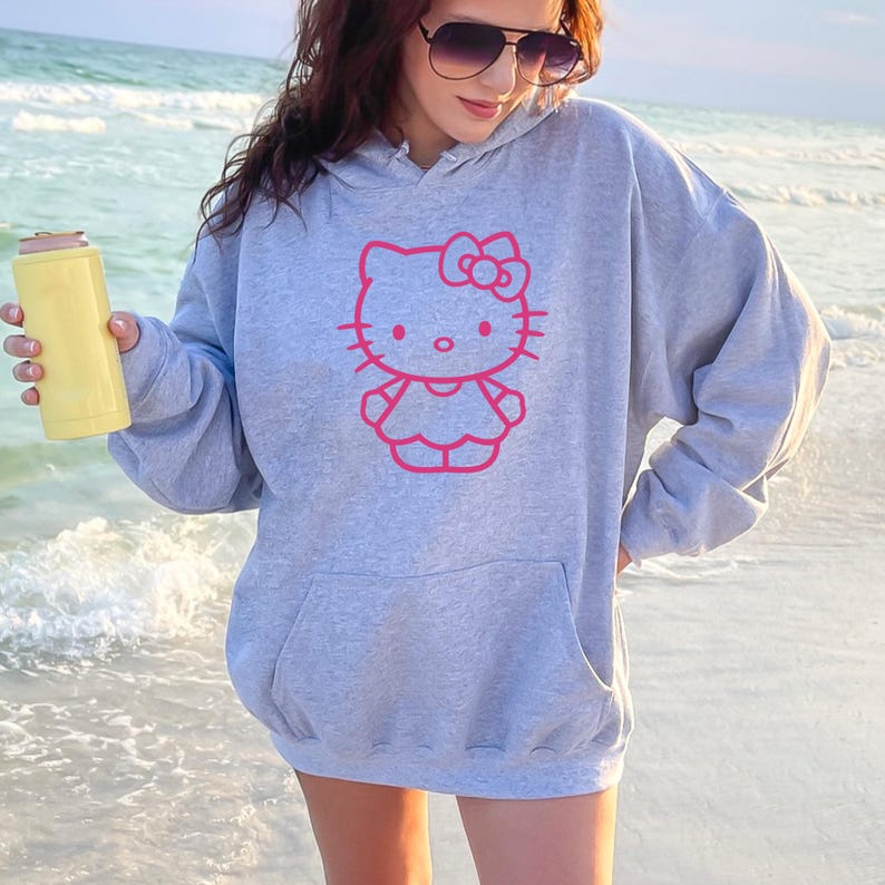 May include: A light gray hoodie featuring a pink outline of Hello Kitty. The hoodie has a front pocket and a drawstring hood. A yellow can cooler is held in the hand. The background is a beach with ocean waves.