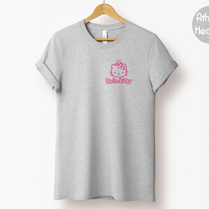 May include: A heather gray t-shirt with a pink Hello Kitty graphic on the front. The graphic includes the Hello Kitty character and the text "Hello Kitty" in a pink font. The shirt is hanging on a wooden hanger.