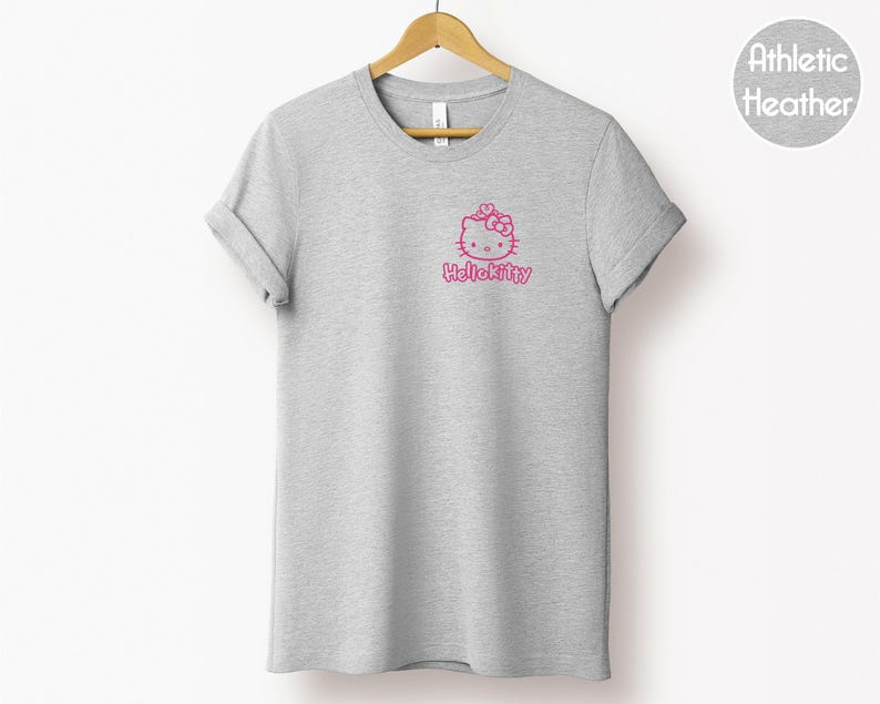May include: A heather gray t-shirt with a pink Hello Kitty graphic on the front. The graphic includes the Hello Kitty character and the text "Hello Kitty" in a pink font. The shirt is hanging on a wooden hanger.