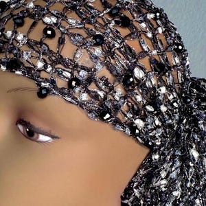 May include: A close-up of a black and silver head covering. The head covering is made of a woven material with black beads and silver accents. The head covering is on a mannequin head.