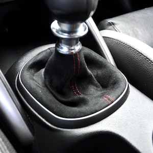 Può includere: Una cuffia del cambio in camoscio nero con cuciture rosse a contrasto, abbinata a un pomello del cambio in metallo argentato e nero, posizionata nella console centrale di un abitacolo con sedili in pelle nera visibili ai lati.