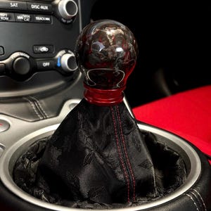 May include: A spherical, dark red and black translucent gear shift knob with a metallic red base collar, attached to a black fabric shift boot featuring a subtle floral pattern and a single vertical line of red stitching in the center.