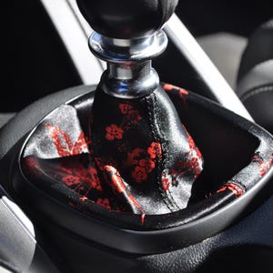 May include: A car gear shift knob with a metallic silver base and a black fabric boot featuring a repeating red floral pattern. The gear shifter is positioned within a black plastic console frame, showing a smooth, dark, and textured finish.