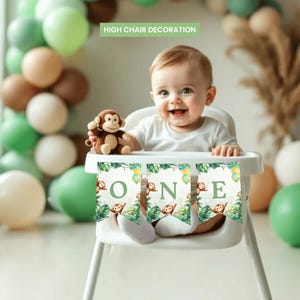 Peut inclure: Un bébé souriant est assis dans une chaise haute décorée d'une bannière avec le mot "ONE" en lettres vertes. La bannière a un thème jungle avec des singes et des feuilles. Un singe en peluche est sur le plateau. Des ballons verts, marron et blancs sont en arrière-plan.