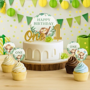 Op de afbeelding: Een witte taart met een gouden nummer één en het woord "One" in gouden letters. De taart is versierd met een apen thema en de woorden "Happy Birthday". Cupcakes met apen toppers staan voor de taart.