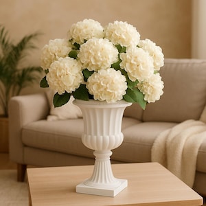 Peut inclure: Un vase urne blanc cannelé rempli de fleurs d'hortensia crème. Le vase est posé sur une table en bois de couleur claire. Les fleurs ont des feuilles vertes et sont disposées en une présentation pleine et luxuriante. L'arrière-plan comprend un canapé beige.