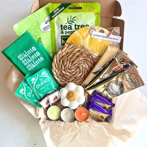 Può includere: Una confezione regalo piena di articoli per la cura di sé. Include maschere per il viso verdi, bustine di tè, un sottobicchiere intrecciato, bustine di caffè, candele e un sapone a forma di fiore. È visibile anche una calza verde con la scritta "Sláinte".