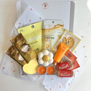 Può includere: Una scatola regalo bianca contenente due bustine di caffè Mokate Gold, una maschera in tessuto gialla PS, una maschera viso Bioaqua, patch dorati per occhi, una candela a forma di fiore, due lumini arancioni, una crema mani e due pacchetti di biscotti Biscoff.
