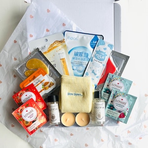 May include: A gift box filled with self-care items, including face masks, tea sachets, candles, and small bottles of shampoo and shower gel. The box contains a yellow washcloth with the words "Slow Down" printed on it. The items are arranged on white tissue paper with pink hearts.