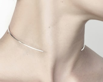 Può includere: Un girocollo in argento con un design semplice e curvo. Il girocollo è indossato da una persona con i capelli corti.