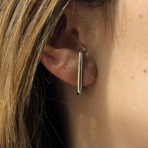 Puede incluir: Un pendiente de aro de plata con un diseño rectangular simple. El aro se lleva en la parte superior de la oreja.