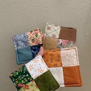 May include: Three square fabric coasters featuring patchwork designs. One coaster displays blue, red, and white floral and checkered patterns. Another shows brown, beige, and floral prints. The third features green, white, and orange floral motifs.