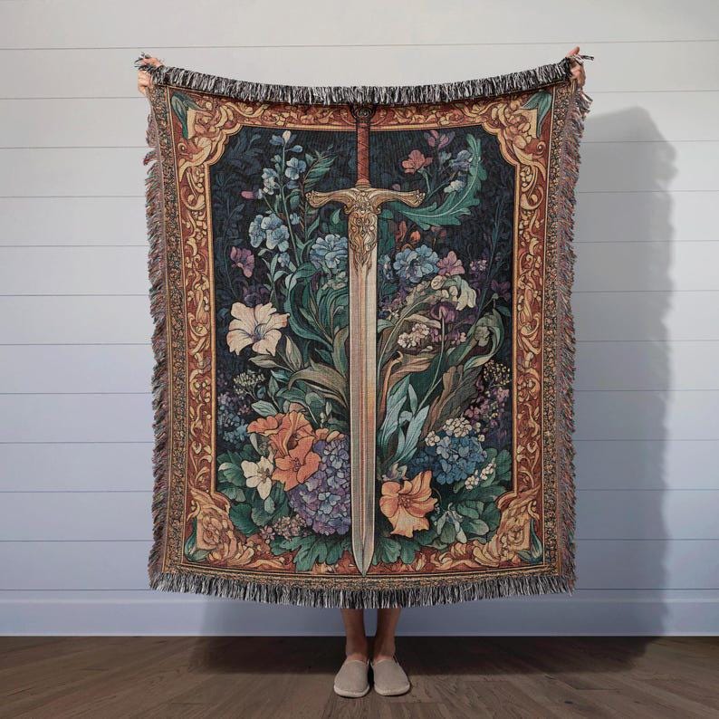 Puede incluir: Una manta de tapiz tejido con un dise&ntilde;o central de espada rodeada de flores coloridas. La manta tiene un fondo oscuro y un borde decorativo en tonos marrones, dorados y verdes. Los bordes est&aacute;n deshilachados.