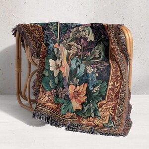 Puede incluir: Una manta de tapiz floral drapeada sobre una silla de mimbre. La manta presenta un fondo oscuro con flores coloridas en tonos naranja, verde, azul y morado. Los bordes tienen flecos.