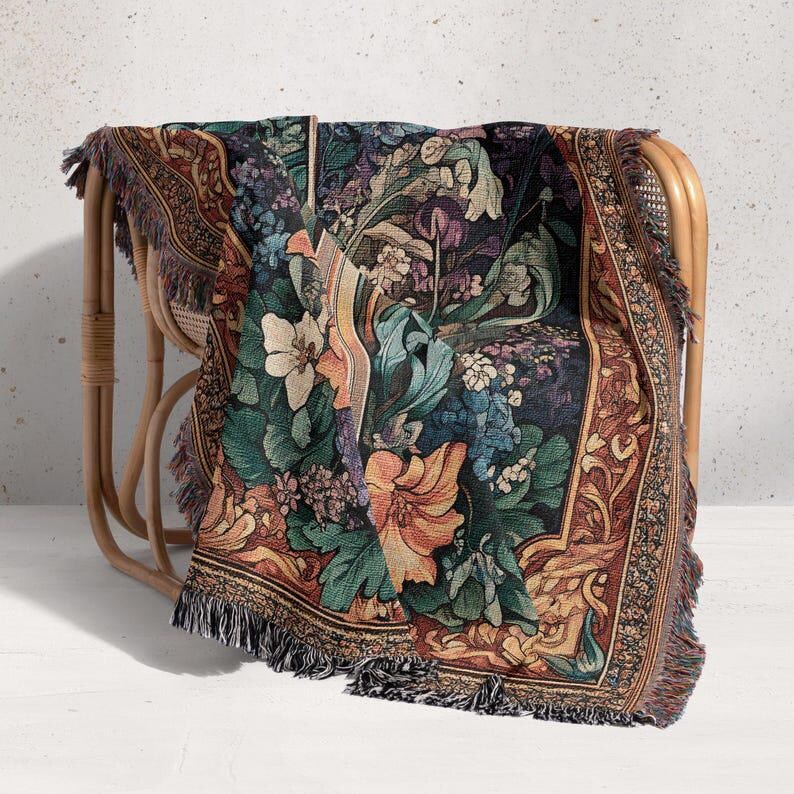 Puede incluir: Una manta de tapiz floral drapeada sobre una silla de mimbre. La manta presenta un fondo oscuro con flores coloridas en tonos naranja, verde, azul y morado. Los bordes tienen flecos.