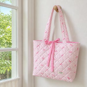 May include: A quilted tote bag featuring a pink and white gingham pattern with small pink bow motifs. The bag has two shoulder straps, a pink ribbon tie closure at the center, and a small rectangular label with the text LEM at the bottom right.