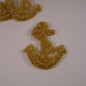 May include: A gold beaded anchor charm. The anchor is made of small, gold-colored beads that are sewn together to create the shape of an anchor.