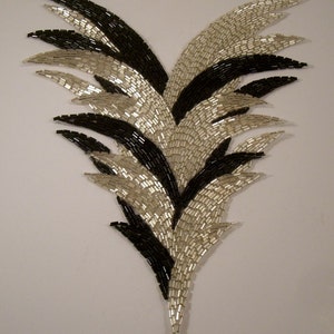 May include: A black and silver beaded design in the shape of a stylized feather. The design is made up of small, rectangular beads that are arranged in a pattern of overlapping layers.