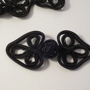 May include: Black fabric Chinese knot closure with a flower design. The knot is made of two pieces that are connected in the middle.
