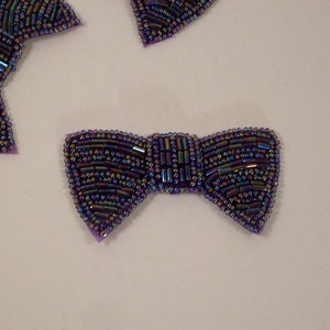 May include: A purple iridescent beaded bow tie. The bow tie is made of small, round beads that are sewn together to create a bow shape. The beads are iridescent, meaning they change color depending on the angle of light.