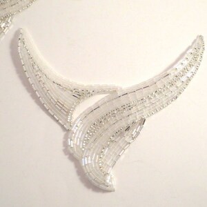 May include: Two white beaded embellishments in the shape of a stylized wave or wing. The embellishments are made of small, white beads sewn together in a flowing pattern.