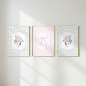 May include: Three rectangular wall art prints in gold frames. The outer two prints feature a blue and green floral pattern with a central pink hydrangea illustration. The middle print displays a pink striped background with a pink ribbon bow.