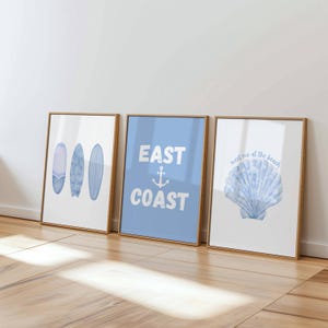 May include: Three framed art prints with a coastal theme. The first print features three blue surfboards. The second print has the words "EAST COAST" with an anchor. The third print shows a blue seashell with the text "meet me at the beach."