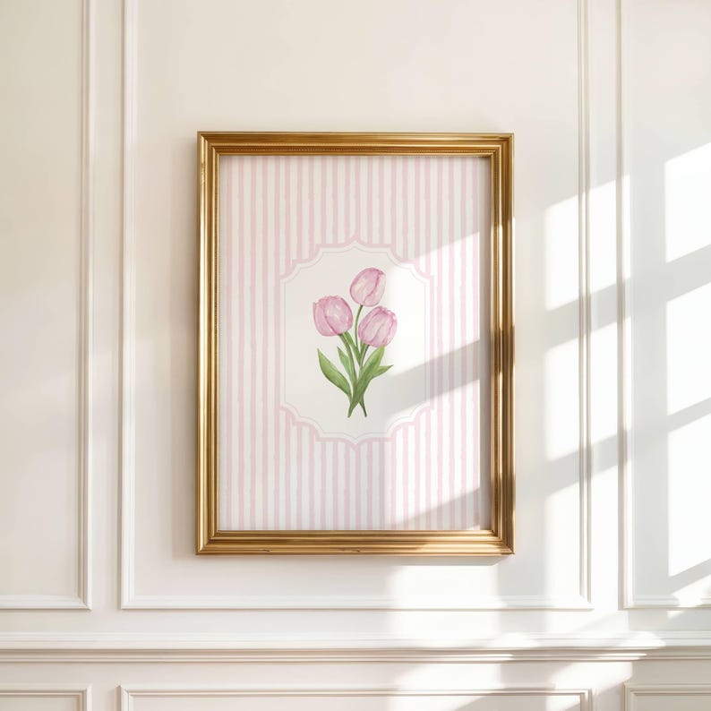 Op de afbeelding: Een ingelijste aquarelprint met drie roze tulpen met groene bladeren. Het kunstwerk staat tegen een roze en witte gestreepte achtergrond in een goudkleurige lijst. De ingelijste kunst hangt aan een witte muur.