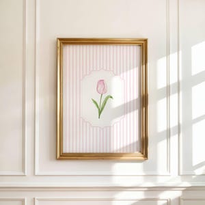 Op de afbeelding: Een ingelijste aquarelprint van een roze tulp. Het kunstwerk staat tegen een roze en wit gestreepte achtergrond, omlijst door een goudkleurige lijst. De ingelijste kunst hangt aan een witte muur.