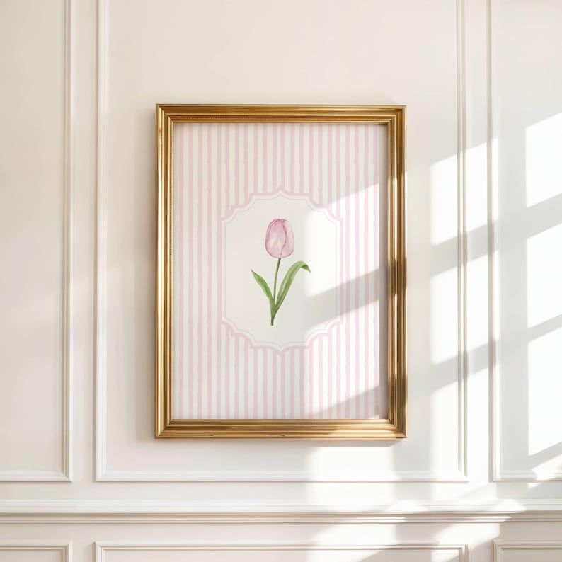 Op de afbeelding: Een ingelijste aquarelprint van een roze tulp. Het kunstwerk staat tegen een roze en wit gestreepte achtergrond, omlijst door een goudkleurige lijst. De ingelijste kunst hangt aan een witte muur.