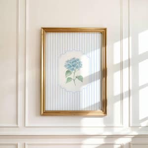 May include: A framed floral print featuring a blue hydrangea blossom. The artwork is set against a backdrop of vertical blue and white stripes, framed in a gold-colored frame. The print is mounted on a white wall.