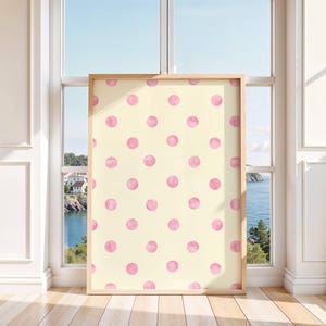 May include: A framed artwork featuring a pattern of pink watercolor polka dots on a cream background. The wooden frame surrounds the artwork, which is displayed in front of a window overlooking a coastal scene.