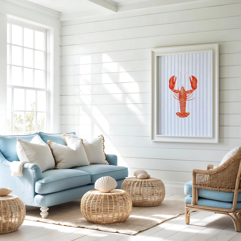 Op de afbeelding: Een woonkamer met kustthema met een lichtblauwe bank, rotan meubels en een ingelijste kunstprint van een rode kreeft op een blauw-wit gestreepte achtergrond. Decoratieve witte kussens en schelpaccenten maken de nautische inrichting compleet.