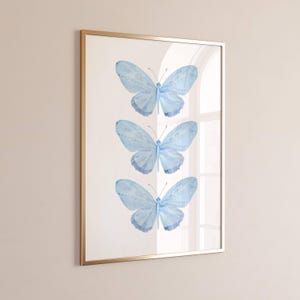 May include: A framed art print featuring three light blue butterflies with intricate wing patterns. The butterflies are arranged vertically on a white background, encased in a gold-colored frame. The artwork is a decorative piece.