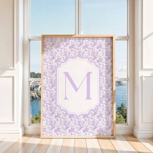 Puede incluir: Un estampado enmarcado con un patrón floral en tonos lavanda y blanco. El estampado tiene un marco de madera clara y un elemento de diseño central con la letra "M" en lavanda. El fondo muestra una vista costera.