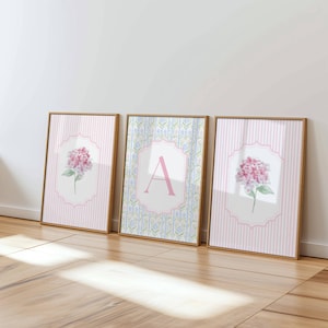 Könnte beinhalten: Drei gerahmte Wandbilder in hellen Holzrahmen. Die beiden äußeren Bilder zeigen rosa Hortensien-Illustrationen auf hellrosa gestreiftem Hintergrund. Das mittlere Bild zeigt ein großes rosa A auf einem blau-weißen Blumenmuster.