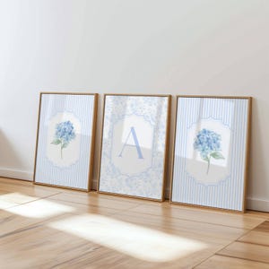 Op de afbeelding: Drie ingelijste wandkunstprints met lichte houten lijsten. De buitenste twee prints tonen blauwe hortensiabloemen op een lichtblauwe en witte gestreepte achtergrond. De middelste print toont een grote blauwe letter A op een bloemmotief achtergrond.
